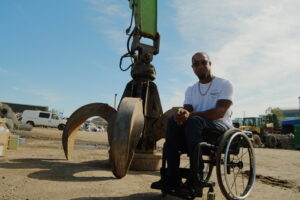 Anthony is sitting in front of the magnet and claws attached to the arm of the crane that crushed him 15 years ago